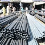 Electrical conduit pipes stacked in a vibrant Pakistani market with workers in traditional attire handling PVC and metal conduits under bright sunlight.